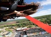 A person with long hair rides an inverted roller coaster, smiling with arms pressed to the harness, with a view of trees and buildings below on a sunny day.