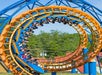 A roller coaster car filled with people goes upside down through a vertical loop on a yellow and blue track, with trees and rooftops in the background.