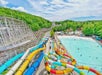 A water park with colorful water slides, a large wave pool with people swimming, and a wooden roller coaster surrounded by green trees under a partly cloudy sky.