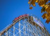 A red roller coaster train ascends the peak of a wooden coaster structure under a clear blue sky, with autumn leaves partially visible in the corner.