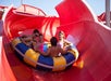 Four people in swimsuits ride together on a large yellow and blue inflatable raft down a red waterslide on a sunny day.
