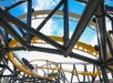 A roller coaster with yellow and white tracks curves through a framework of black supports, carrying passengers under a bright blue sky with scattered clouds.