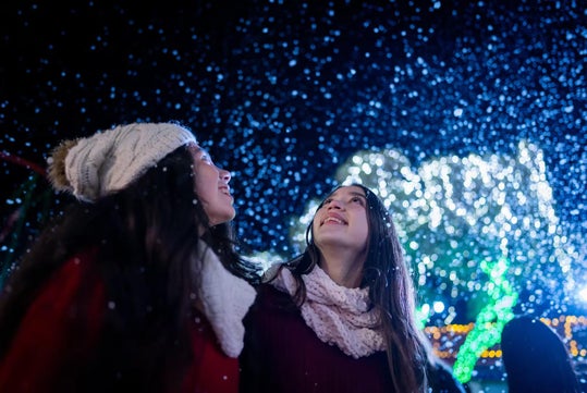 Two people wearing winter clothes look up and smile as artificial snow falls at night, with bright holiday lights in the background.