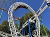 A roller coaster train moves upside down through a vertical loop against a clear blue sky, surrounded by trees and metal supports.
