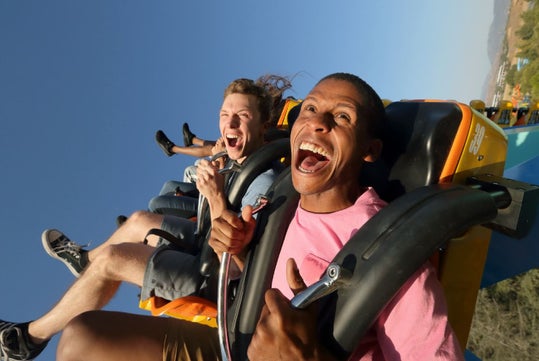 Two people riding a roller coaster in open seats, gripping safety bars and smiling with excitement against a clear blue sky.