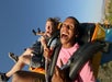 Two people riding a roller coaster in open seats, gripping safety bars and smiling with excitement against a clear blue sky.