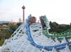 Two roller coasters with blue and green tracks race side by side on a steep white wooden structure, with riders’ arms raised and a red observation tower in the background.