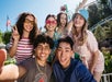 Six young adults pose for a group selfie at an amusement park, smiling at the camera with roller coaster tracks and trees in the background.