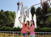 Two young women take a selfie and eat food in front of a fountain, with roller coasters and greenery in the background at an amusement park.