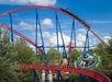 A red and blue roller coaster with riders is surrounded by trees under a partly cloudy sky. A circular green and blue amusement ride is visible nearby.