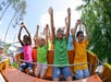 Children in colorful shirts are on an amusement park ride, smiling and raising their arms. Trees and a clear sky are in the background.