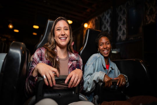 Two women sit side by side in amusement park ride seats, smiling and looking ahead in a dimly lit indoor setting.