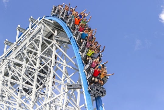 People riding a roller coaster with raised arms, traveling down a steep blue and white track against a clear blue sky.