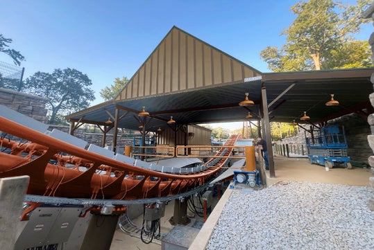 An outdoor roller coaster station with an orange track runs beside a covered waiting area, surrounded by trees and gravel pathways.