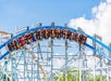 People riding a blue roller coaster on a clear day, with the track curving downward against a blue sky and scattered clouds.