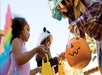 Three children in costumes receive candy from an adult holding a pumpkin bucket during trick-or-treating on Halloween.
