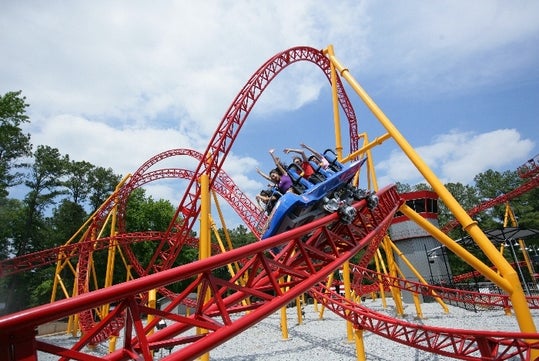Roller coaster with red tracks and yellow supports, carrying several riders with their hands up, set against a clear blue sky and surrounded by trees.
