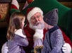 A man dressed as Santa Claus sits on a green chair, listening to two young children, one girl and one boy, as they talk to him.