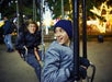 Two boys wearing jackets and beanies sit on swings at an outdoor amusement park at night, with festive lights in the background.
