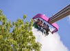 People are seated on a high amusement park ride against a blue sky with clouds. A tree is partially visible in the foreground.