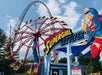 The "SUPERGIRL Sky Flyer" is a vibrant spinning pendulum attraction, featuring a predominantly red ride with yellow and blue accents, matching the Supergirl theme, while in the background, a large roller coaster with a massive red track loops into the sky on a clear, sunny day surrounded by lush green trees at Six Flags St. Louis.
