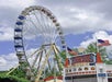 Ferris wheel with colored gondolas stands behind a Justice League-themed ice cream stand. Two American flags are mounted on the stand. Trees and cloudy sky in the background.