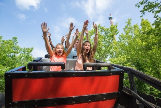 People riding a red and black roller coaster with their hands up, surrounded by trees and blue sky.
