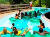 A lazy river full of people floating on clear green tubes while laughing and having a good time on a sunny day.