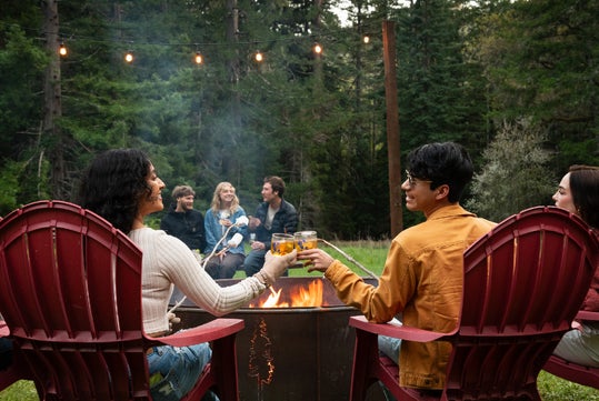 A group of people sit around a fire pit outdoors, toasting drinks and roasting marshmallows, surrounded by trees and string lights.