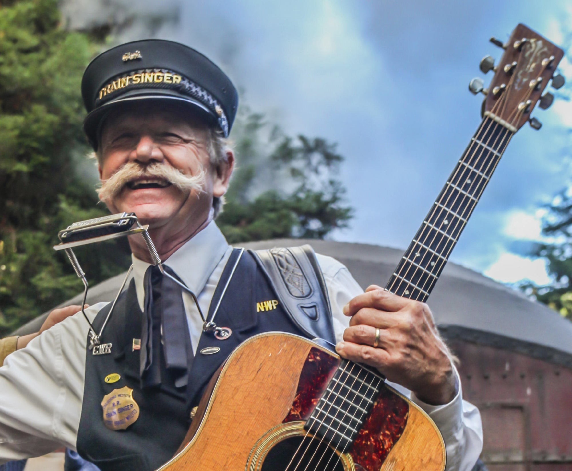 An older man dressed as a train conductor plays a guitar and harmonica outdoors, smiling, with trees and a steam train visible in the background.