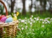 A wicker basket filled with colorful eggs sits on grass amidst small pink and white flowers.