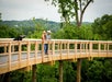Two people wearing hats stand on a wooden boardwalk surrounded by lush green trees, overlooking a scenic landscape.
