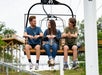 Three young people sit on a ski lift chair outdoors, talking and smiling, with trees and wooden structures visible in the background.