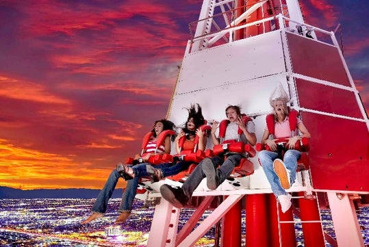 Four people ride an amusement park thrill ride high above a city at sunset, with their hair and clothes blown by the wind.