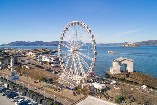 A large Ferris wheel stands near the waterfront with piers, boats, and distant hills, including the Golden Gate Bridge and Alcatraz Island, visible under a clear blue sky.
