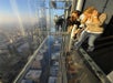 Three people stand on a glass observation ledge high above a city, looking down at the buildings below through the transparent floor.