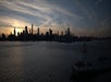 City skyline at sunset with silhouetted skyscrapers, reflecting over the water, and a small lighthouse in the foreground.
