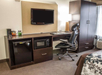 Hotel room workstation with chair, wall-mounted TV, desk, lamp, wardrobe, mini fridge, microwave, coffee maker, and snacks.
