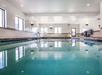 Indoor swimming pool with clear water, surrounded by white walls and large windows. Pool chairs and a door are visible in the background.