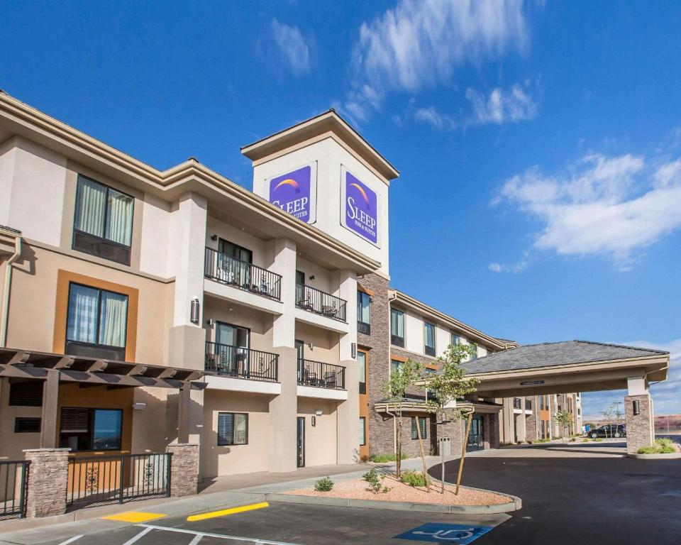 Exterior of a three-story hotel with balconies and a large sign reading "Sleep." The building has beige and brown tones, and the entrance features a covered walkway. Clear blue sky above.