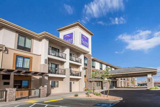 Exterior of a three-story hotel with balconies and a large sign reading "Sleep." The building has beige and brown tones, and the entrance features a covered walkway. Clear blue sky above.