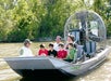 A group of people, including children and adults, sit in an airboat on a river, with a guide at the front and trees in the background.