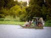 An airboat carrying several people travels on a calm river, surrounded by lush green trees and vegetation.