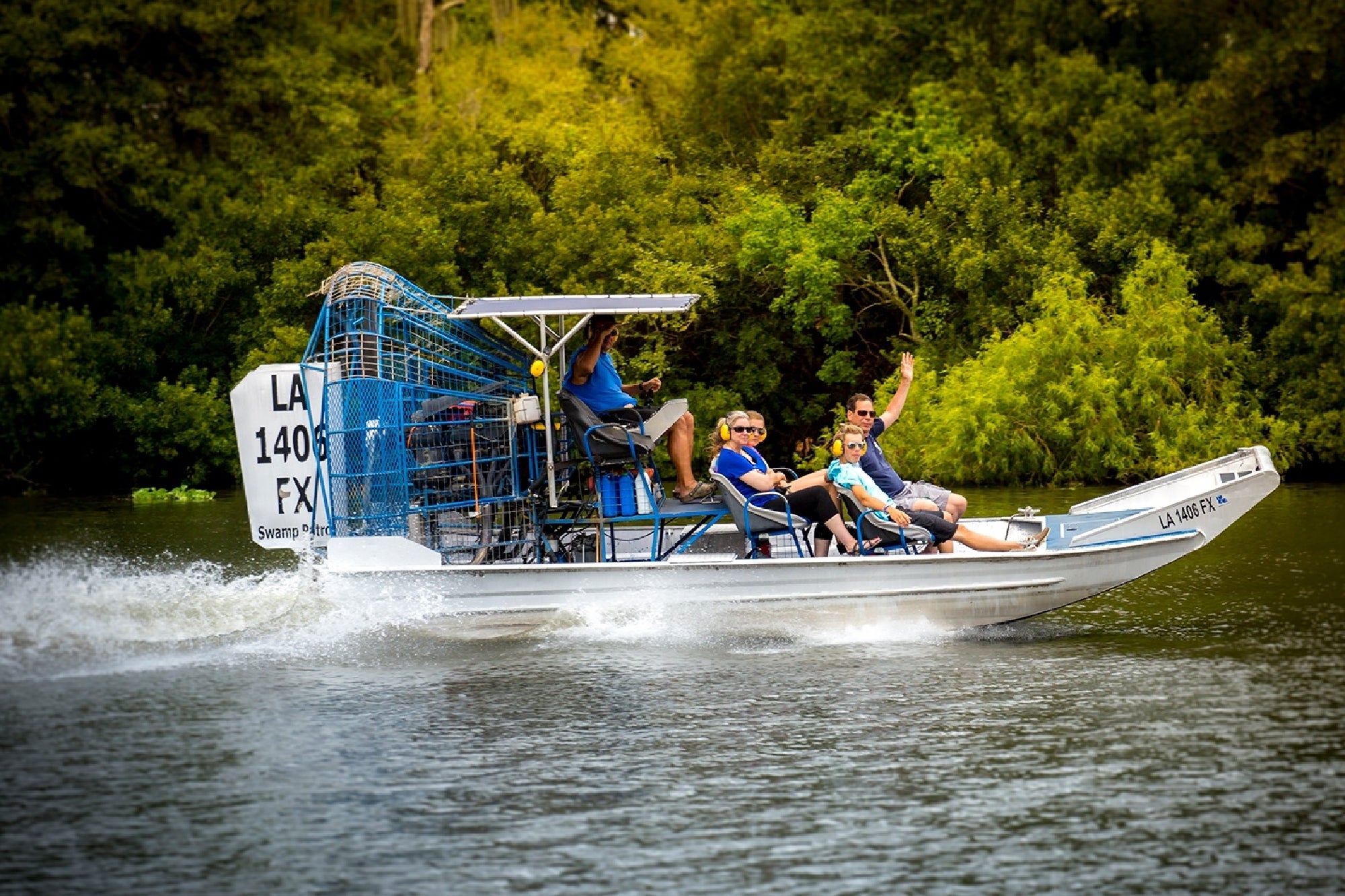 Four people ride an airboat speeding across the water near dense green trees. Two passengers are waving, and the boat creates a visible wake.
