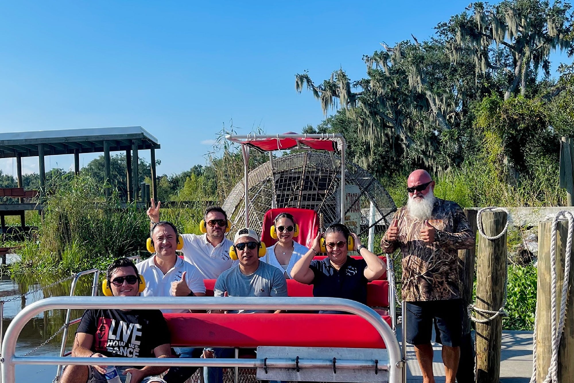 Seven people sit and pose for a photo on an airboat docked by a wooden pier with trees and greenery in the background on a sunny day.