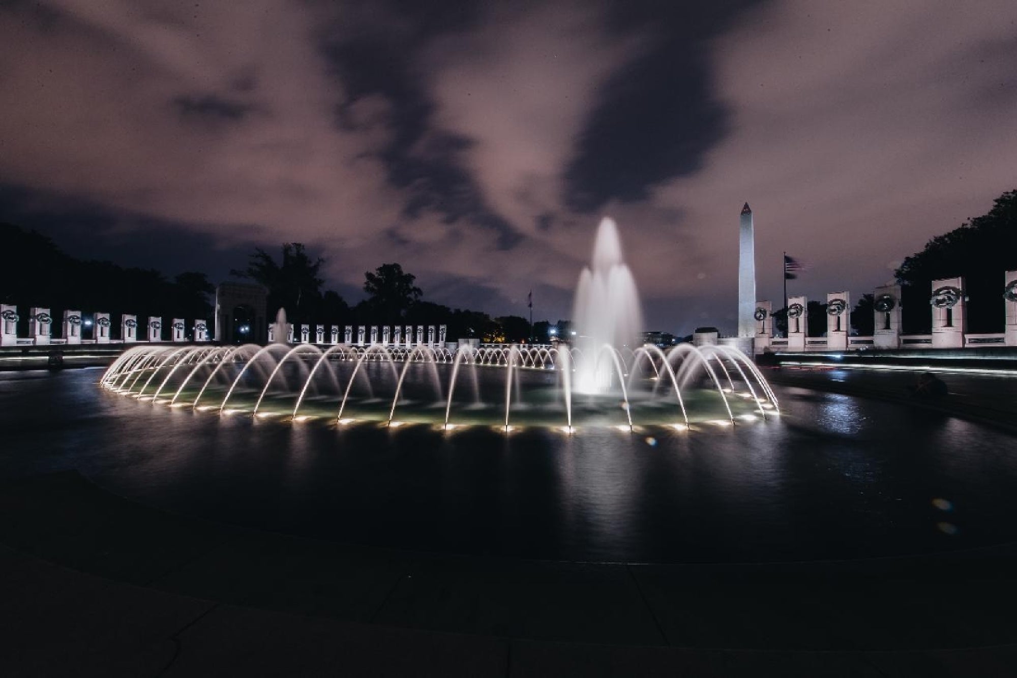 Illuminated WWII Memorial with Washington Monument in the distance.
