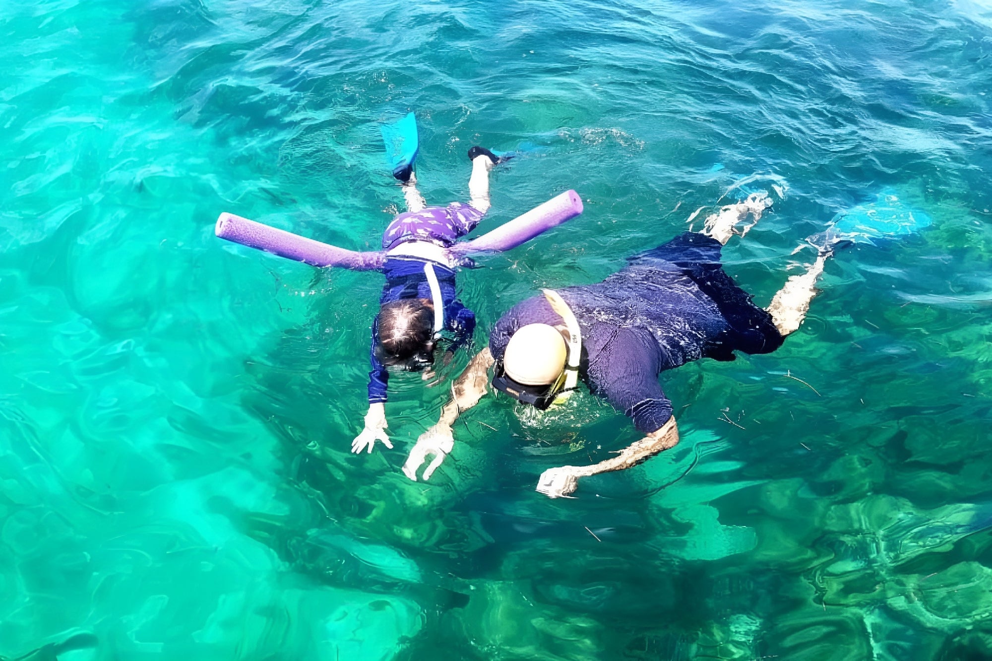 An adult and a child wearing snorkel gear and swim fins float face-down in clear turquoise water; the child uses a purple pool noodle for support.