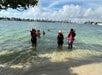 A group of people wearing life jackets stand and swim in shallow, clear water near a shoreline, with a city skyline visible in the background under a partly cloudy sky.