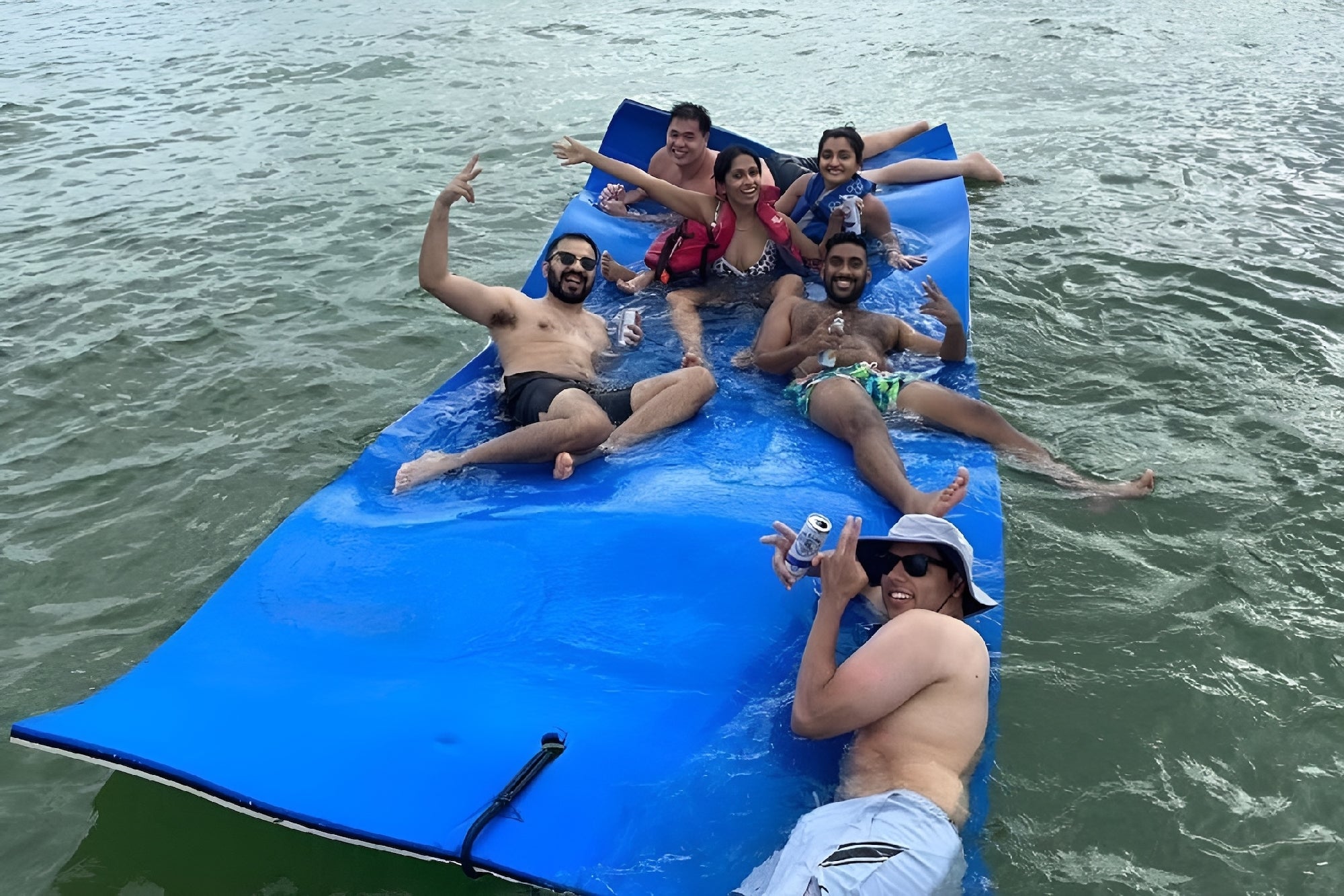 Six people relax on a blue floating mat in the water, some holding drinks and making hand gestures, with a joyful atmosphere.