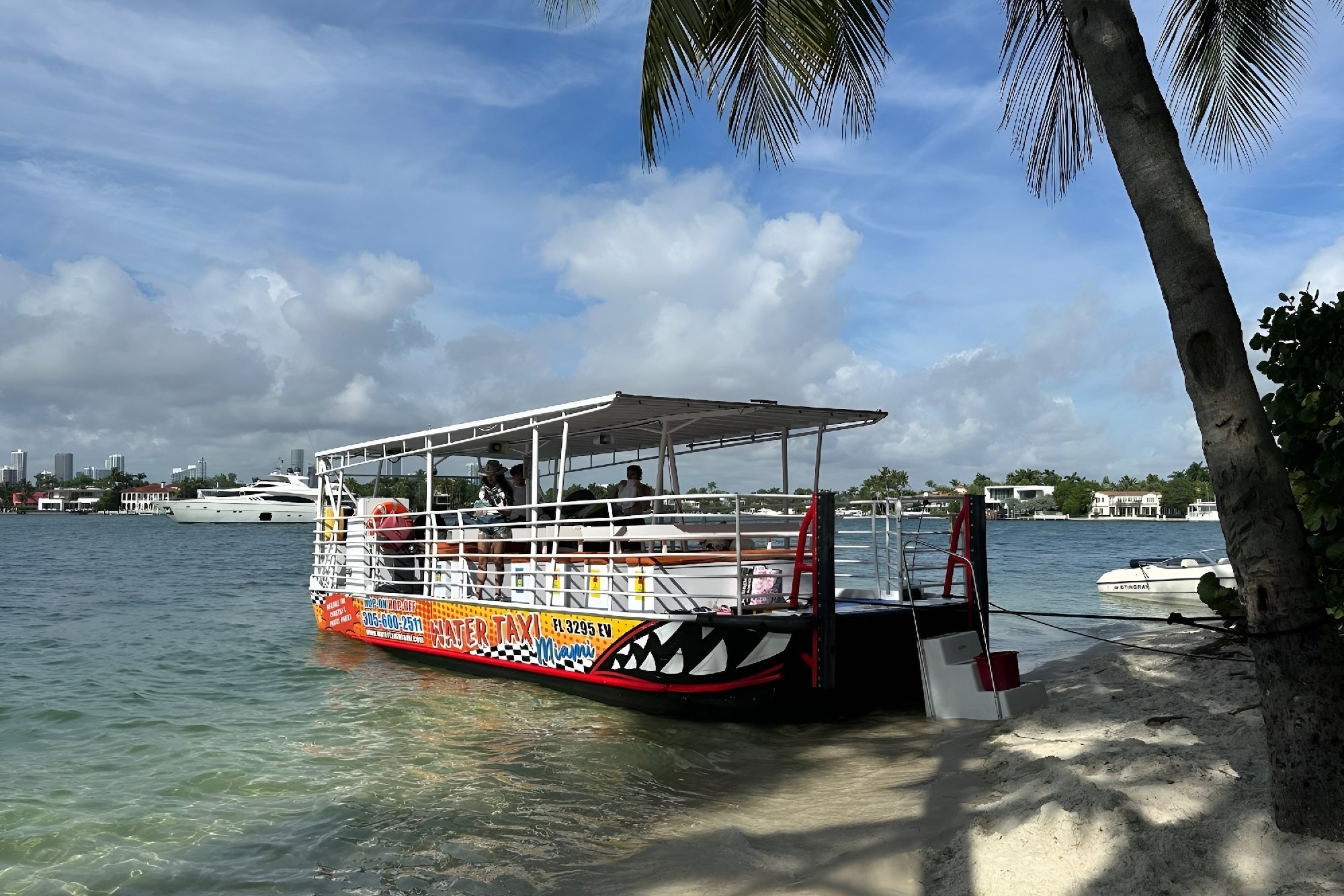 A colorful tour boat is docked near a sandy shore with palm trees, while people are on board and waterfront houses and yachts are visible in the background.